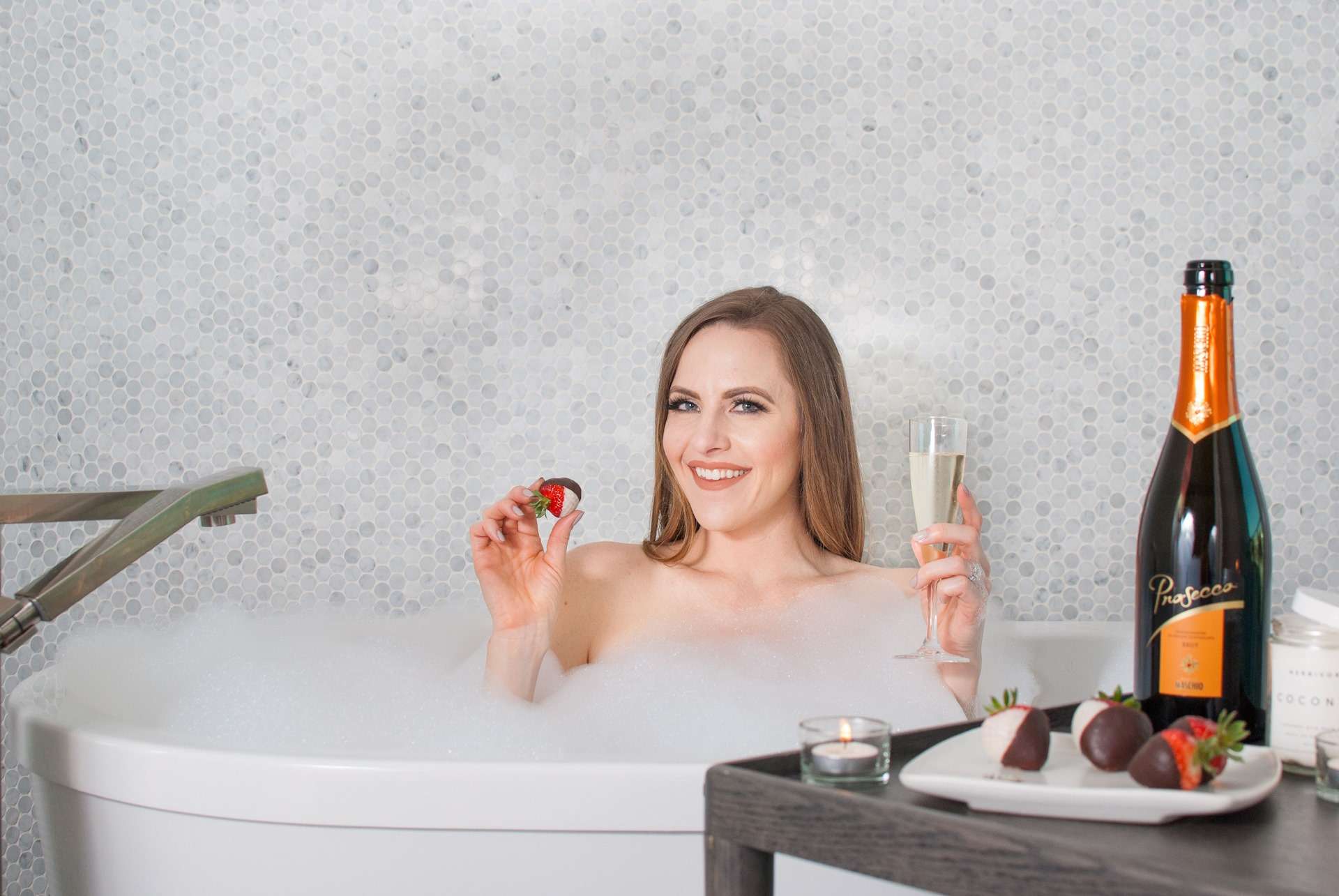 A woman in a bubble bath holding a glass of champagne and chocolate covered strawberries.