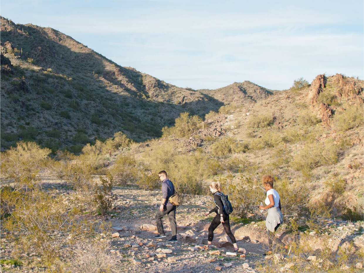 Three individuals trekking a desert trail, mountains in shadow.