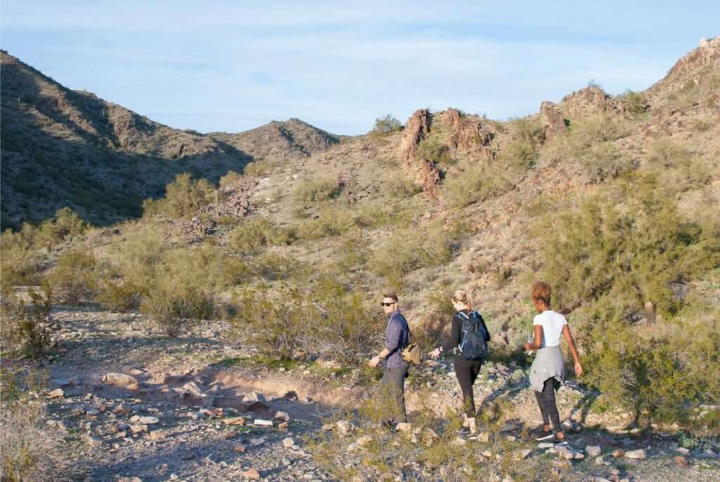 Three people hiking in the desert wearing exercising clothes and backpacks.