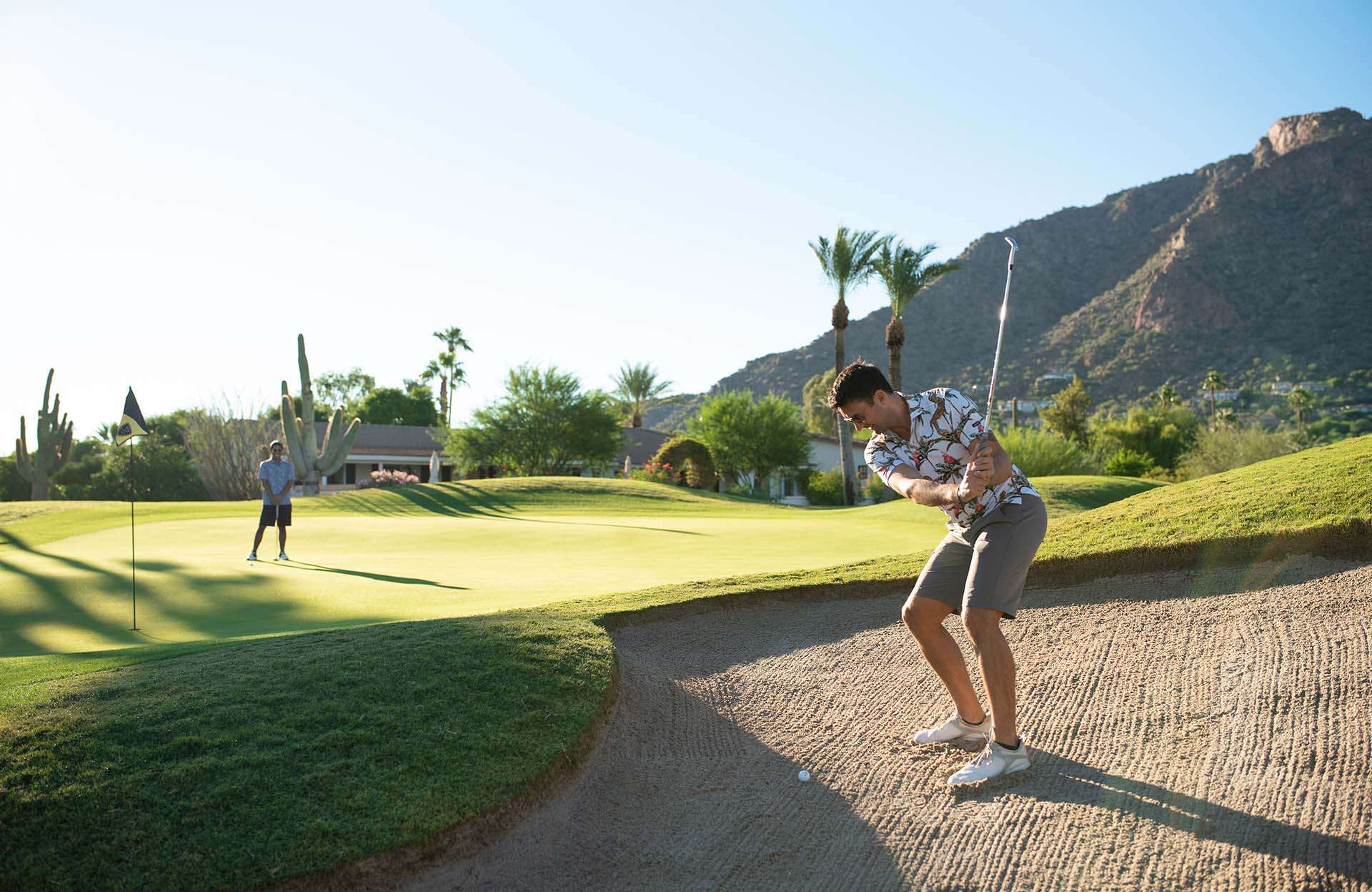 Man getting ready to hit a golf ball with a putt on a golf course with mountains in the back.