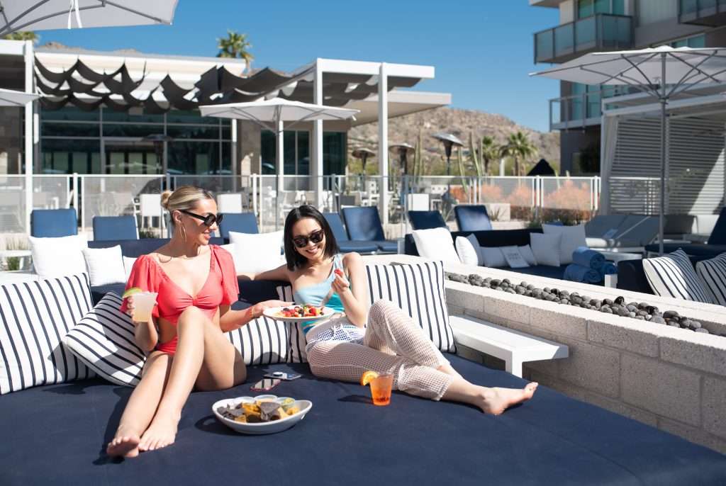 two women sitting on daybed with poolside snacks and cocktails
